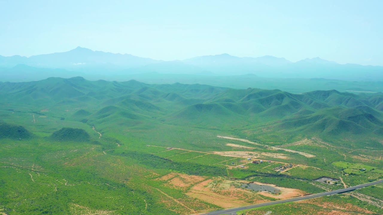 Cerritos Beach, Baja California Sur aerial frame showing a broad green valley, tan field patches, a foreground road curve, layered distant mountains, and no visible people.