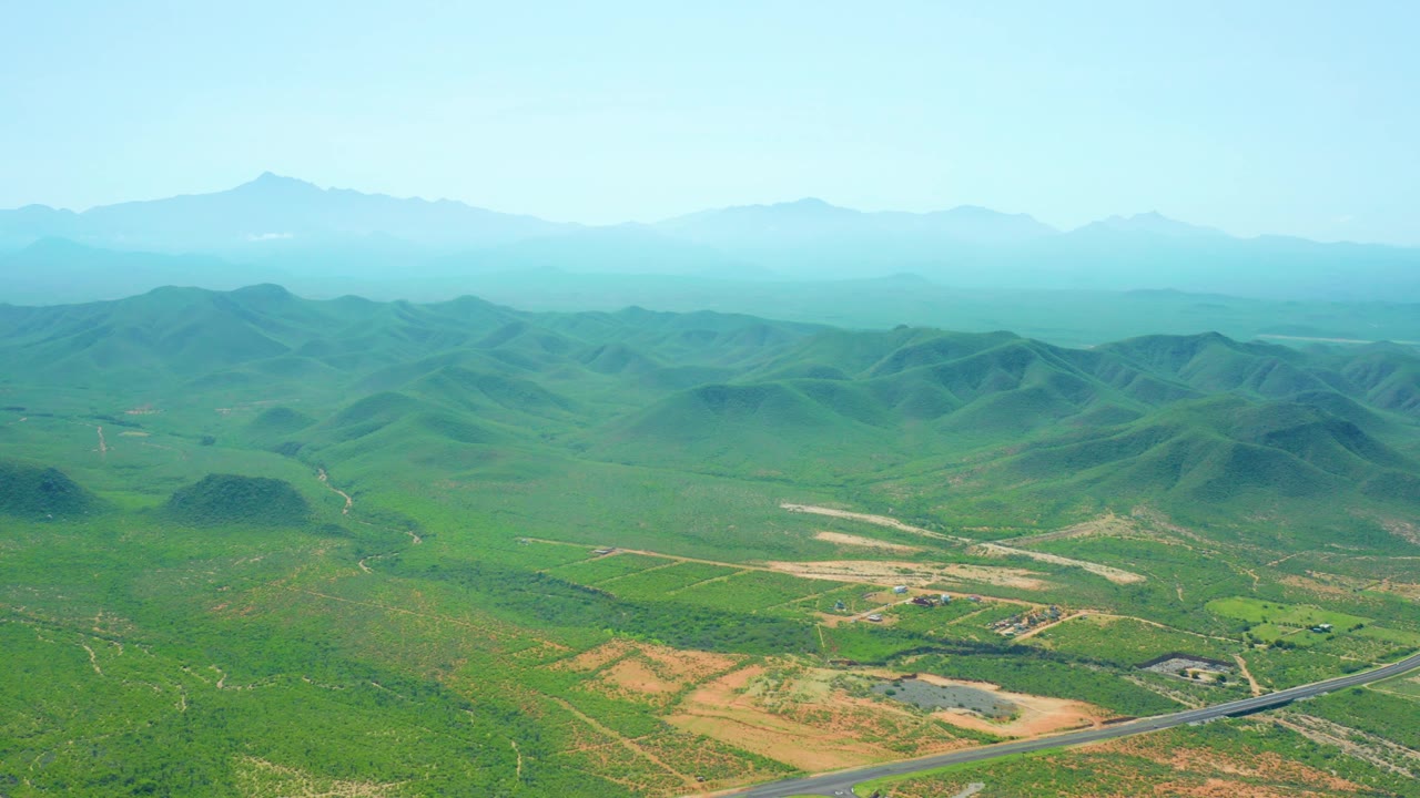 Mid-clip aerial composition over Cerritos Beach, Baja California Sur with rounded green hills across the center, rural fields below, hazy mountains beyond, and no visible people.