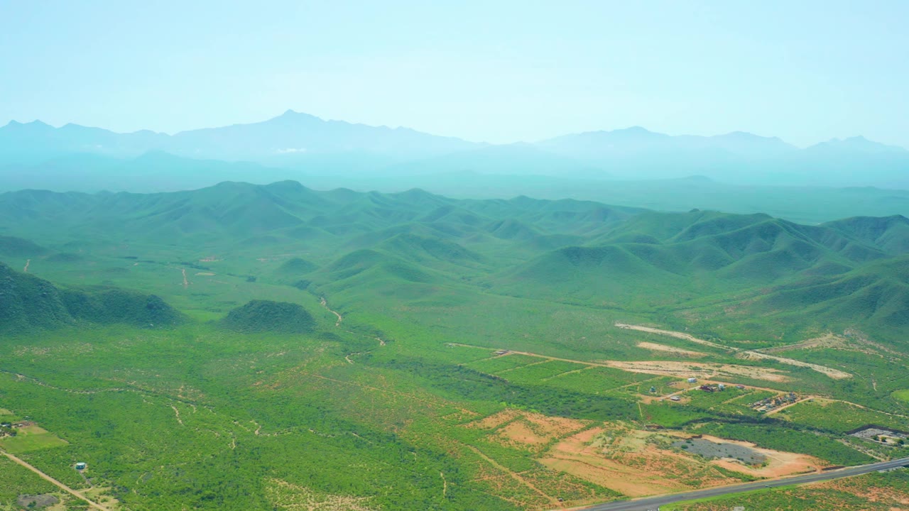Second aerial frame of Cerritos Beach area countryside showing rounded green hills across the middle of the view, pale dirt field areas below, a small road section, distant hazy mountains, and no visible people.