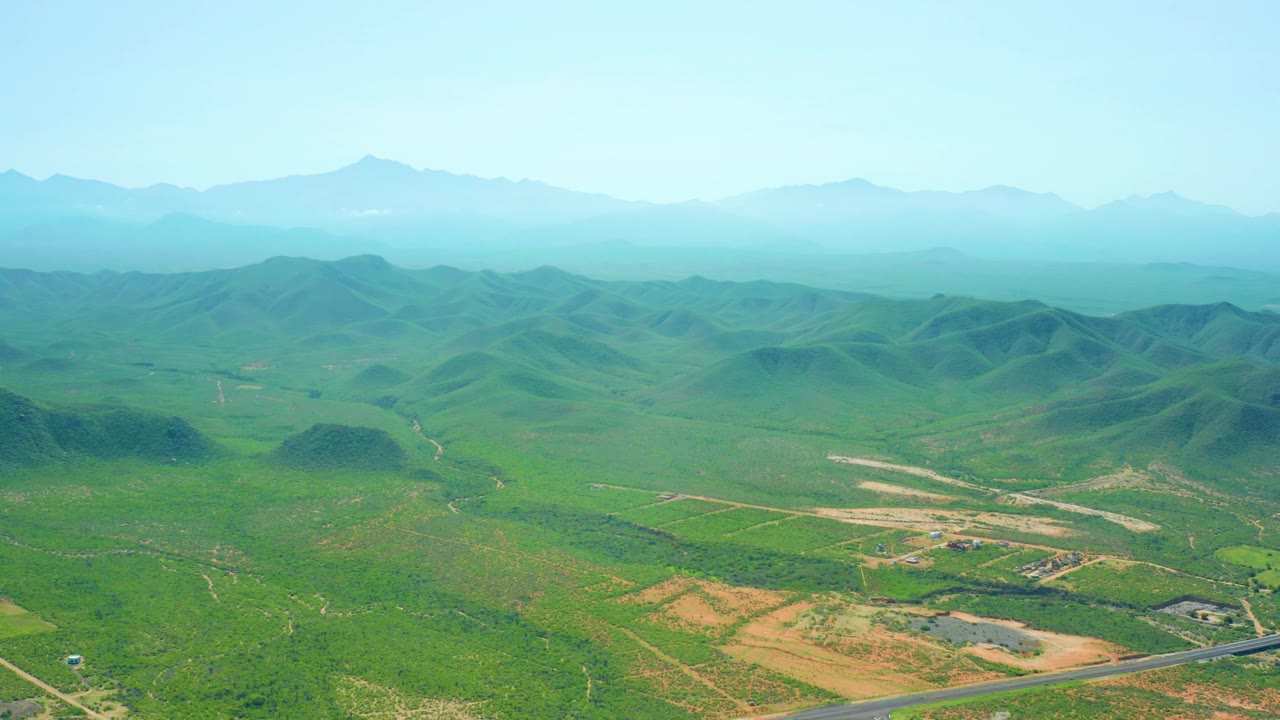 Mid-clip aerial frame over the Cerritos Beach countryside with a high horizon, green rounded hills, pale dirt tracks or fields, distant blue-green mountain layers, and no visible people.