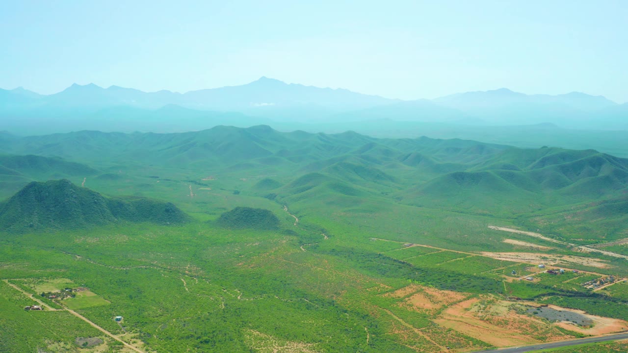 Aerial view across Cerritos Beach, Baja California Sur inland hills with pale dirt tracks, small rural buildings, distant blue ridges, and no visible people.