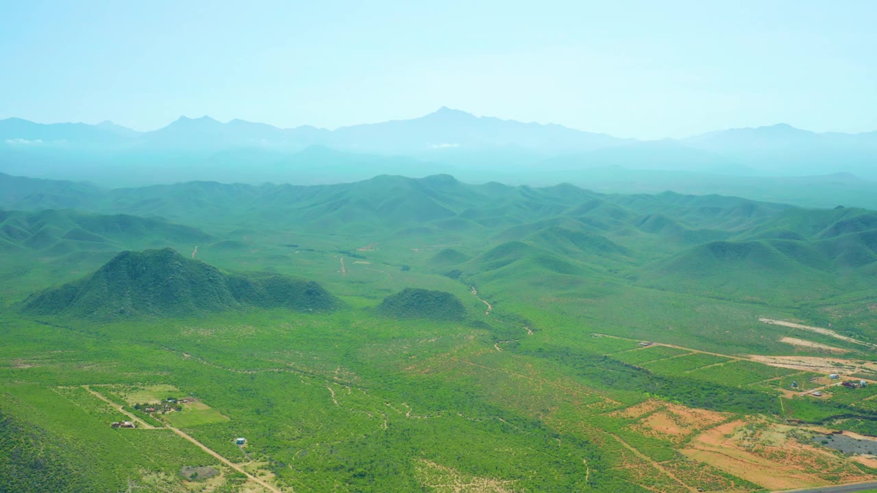 Aerial countryside with rounded green hills, fields, road cuts, and mountain horizon.