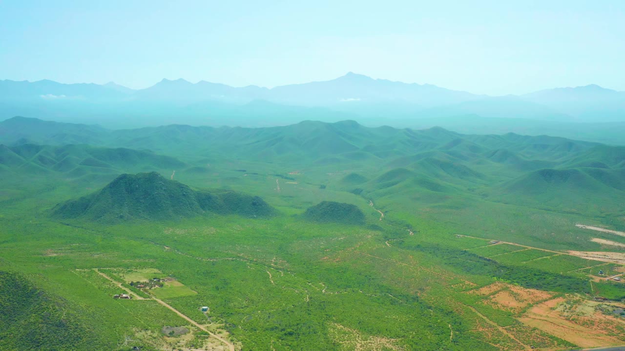 Aerial frame over Cerritos Beach, Baja California Sur, Mexico, showing green farmland, low hills, pale roads, distant mountains, and no visible people.