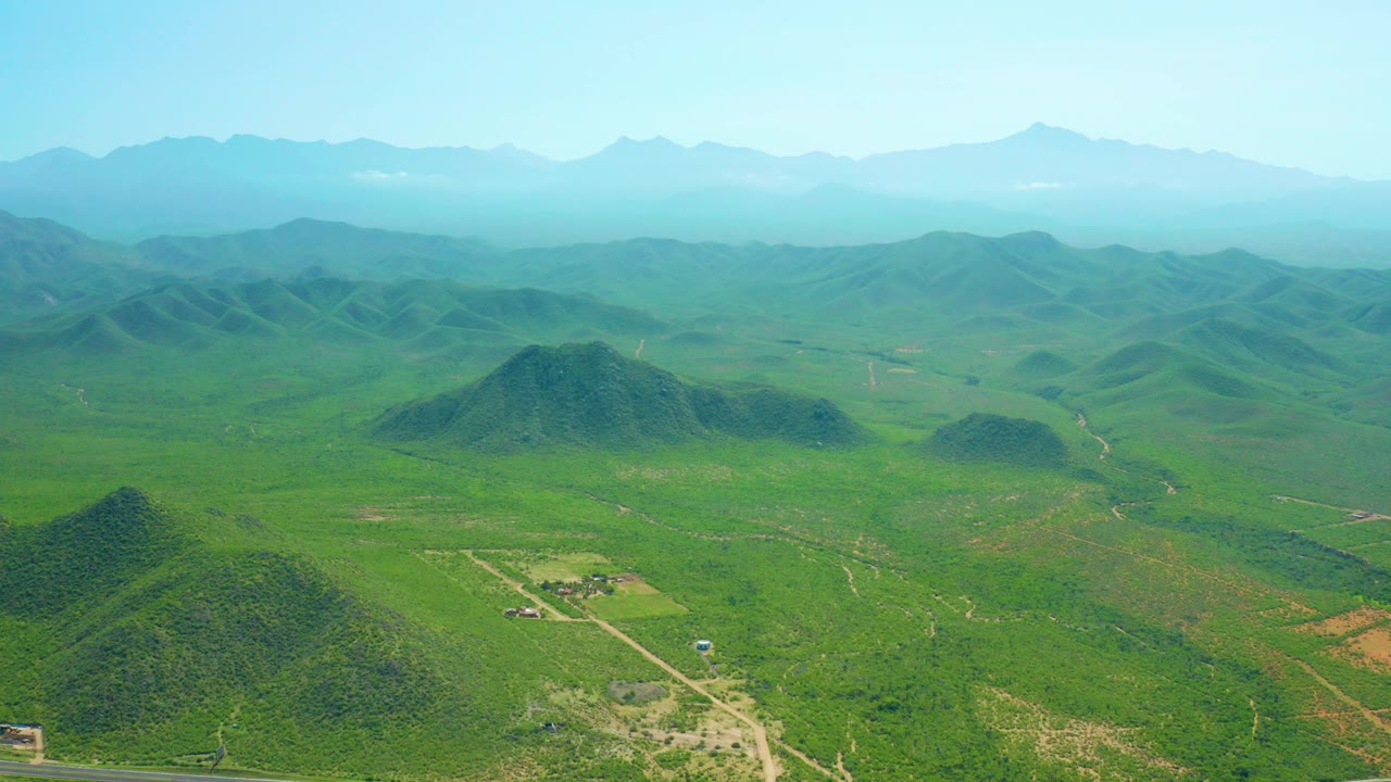 Opening aerial frame over Cerritos Beach countryside with green hills, a paved road along the bottom edge, hazy mountains in the distance, and no visible people.