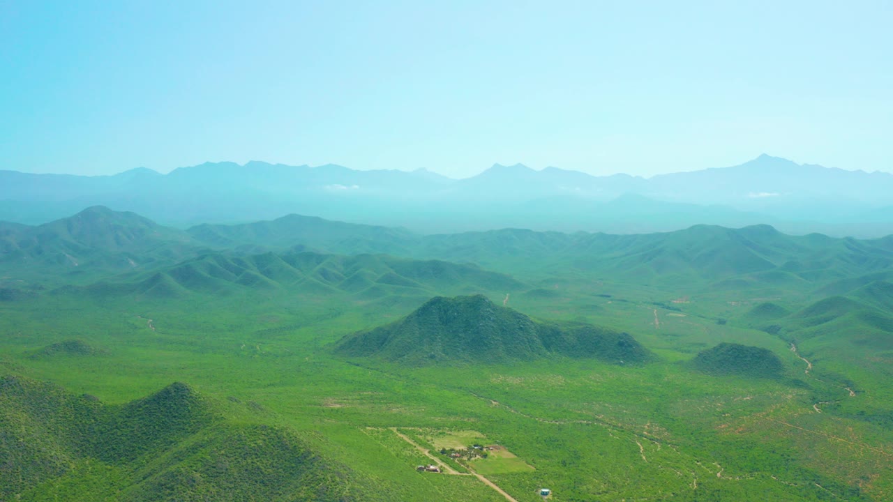 Opening aerial frame over the Cerritos Beach area, showing rounded green hills, a wide valley floor, hazy distant mountains, clear blue sky, and no visible people.