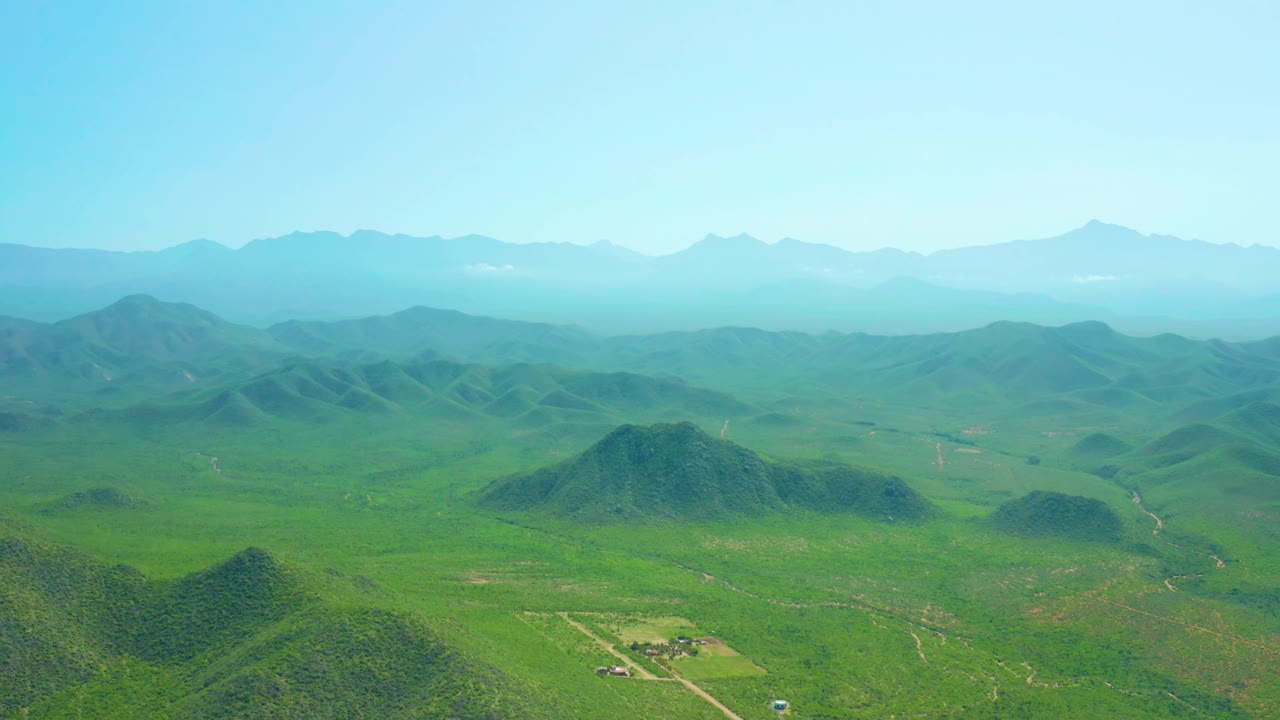 Early frame from the Cerritos Beach BCS clip with a central green hill, open valley floor, pale dirt paths, distant blue mountains, and no visible people.