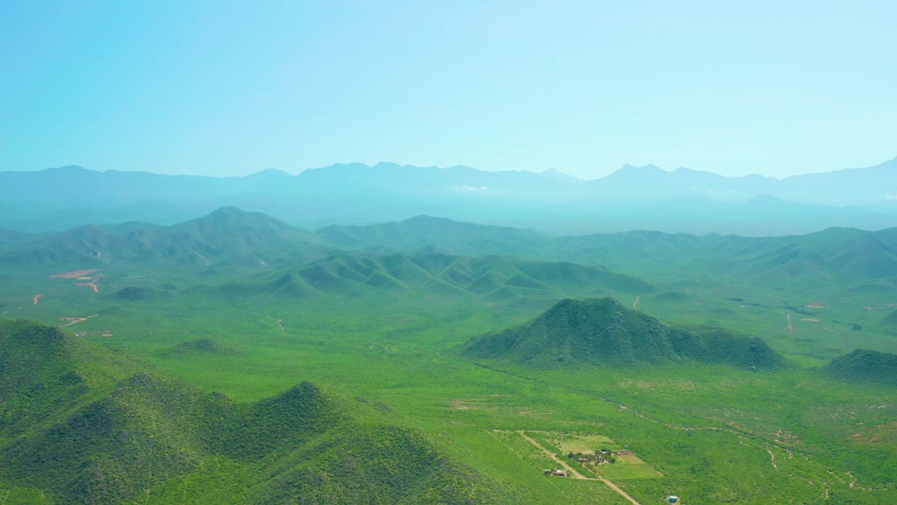 Wide Cerritos Beach, Baja California Sur landscape frame with a low cluster of rural structures below, green hills across the valley, hazy mountains, and no visible people.