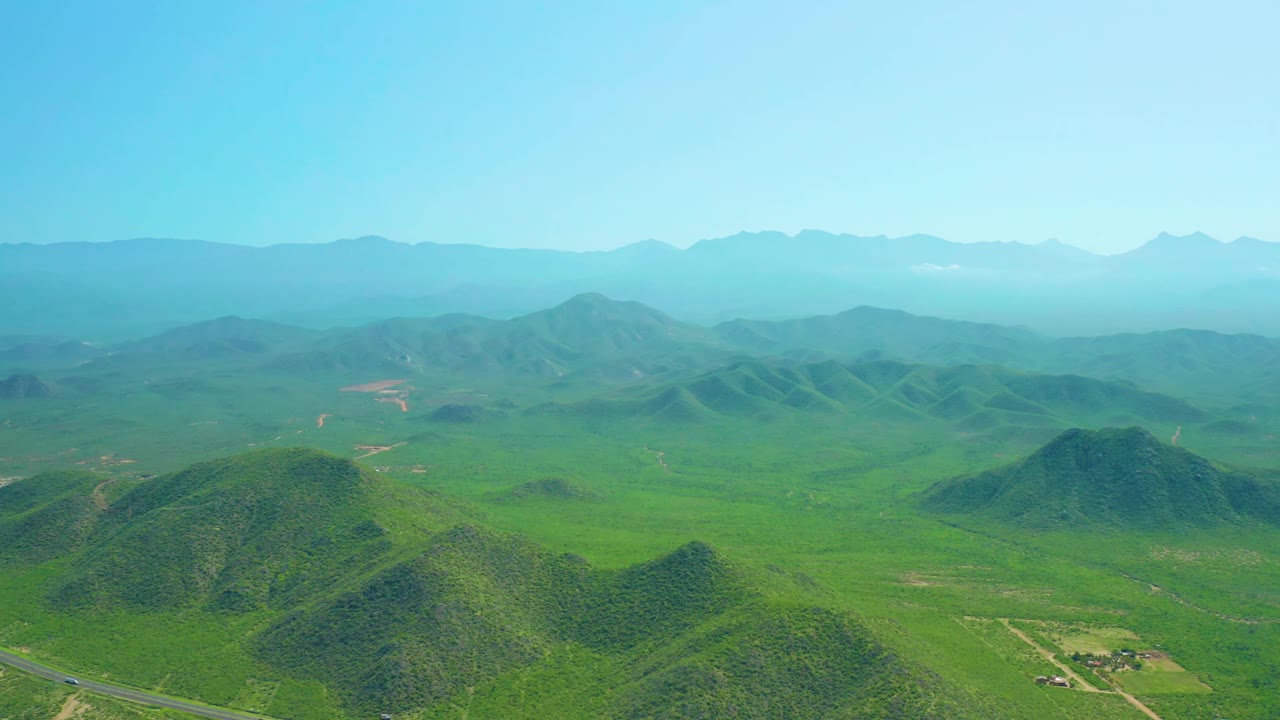 Aerial frame over Cerritos Beach, Baja California Sur showing rolling green hills, a broad valley with field plots, distant mountain ridges, pale blue sky, and no visible people.