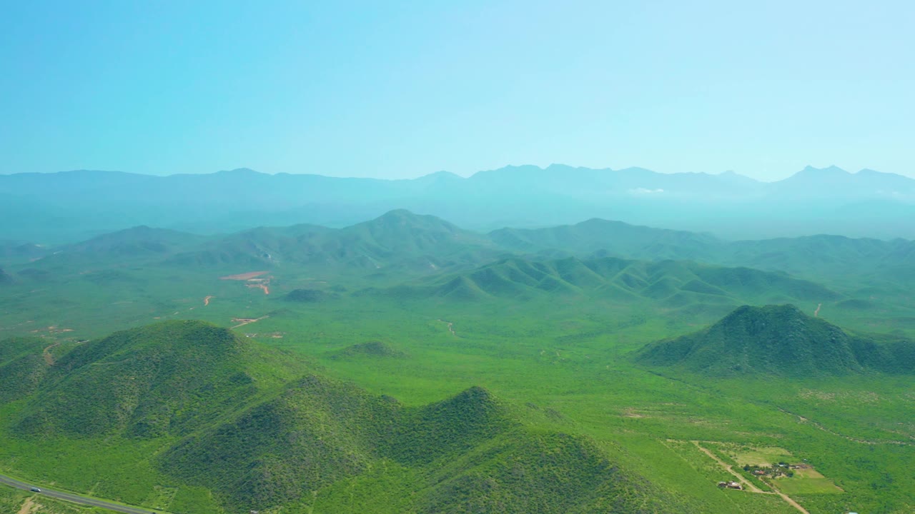 Wide aerial view in Cerritos Beach, Baja California Sur with rounded green hills across the foreground, scattered field patches in the valley, layered blue mountains, clear sky, and no visible people.