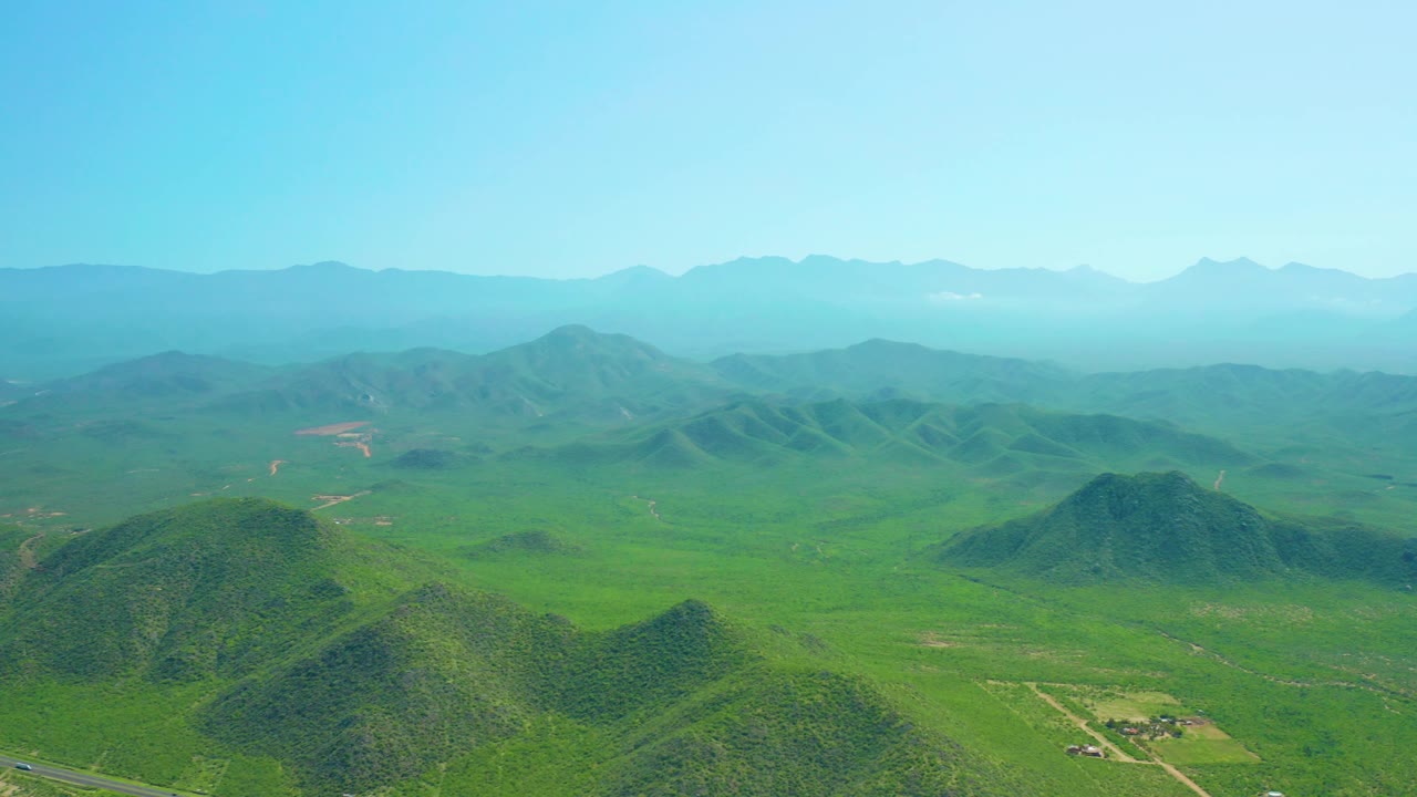 Aerial frame from Cerritos Beach, Baja California Sur showing the road mostly out of the main view, green hills in front, expanded fields to the right, hazy mountains, and no visible people.
