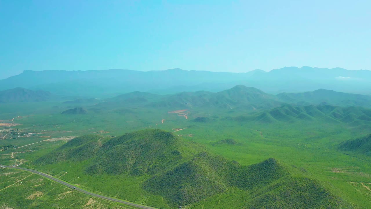 Aerial view in Cerritos Beach, Baja California Sur with low green hills in the foreground, the road more visible along the lower edge, layered mountains, and no visible people.