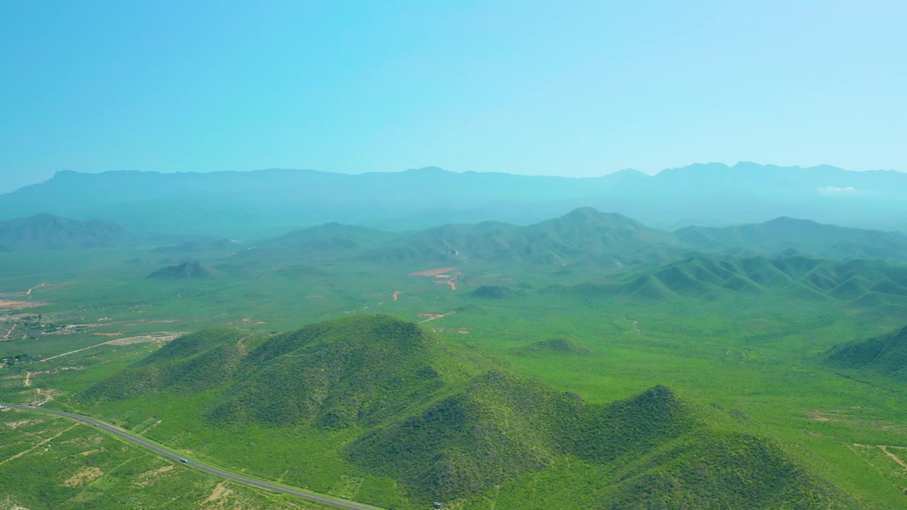 Cerritos Beach, Baja California Sur aerial frame with a broad green valley, clustered rounded hills near center, faint dirt roads, distant mountain silhouettes, and no visible people.