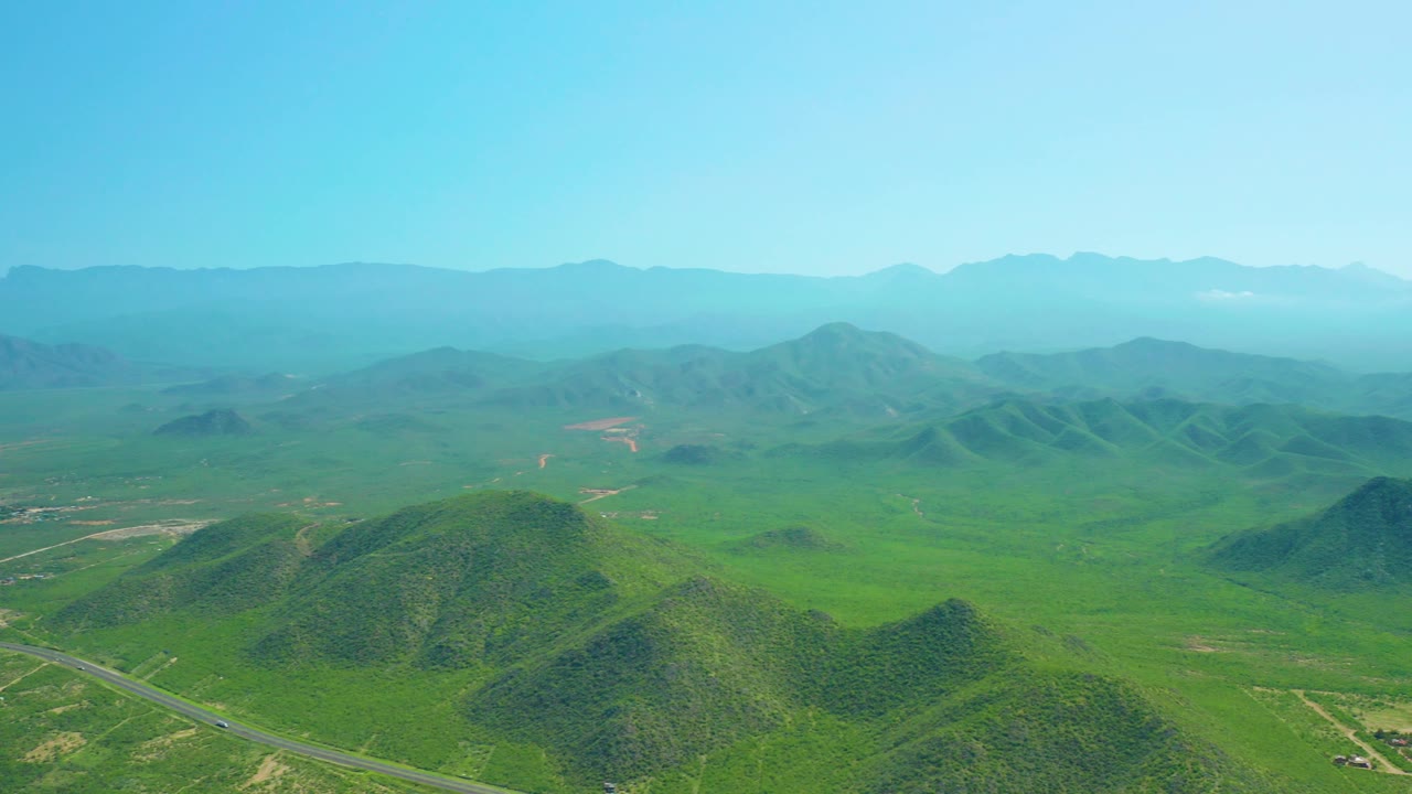 Aerial frame over Cerritos Beach, Baja California Sur showing foreground hills, the paved road near the bottom edge, wide green valley, hazy mountain backdrop, and no visible people.