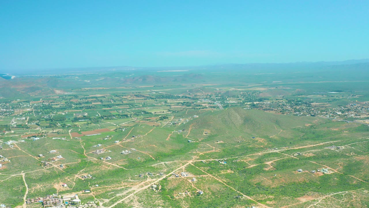 Early frame over Cerritos Beach, Baja California Sur countryside with green and brown fields, pale dirt tracks, scattered buildings, rounded hills, distant mountains, and no visible people.