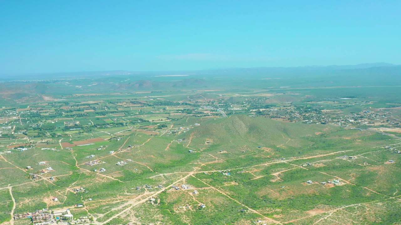 Wide aerial landscape with rural fields, dirt tracks, hills, and a small settlement.
