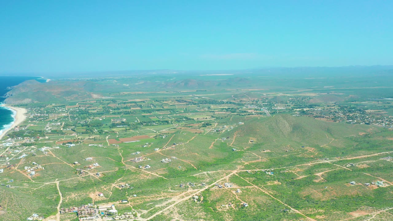 High aerial view over Cerritos Beach, Baja California Sur showing green fields, pale dirt roads, low hills, distant surf, and clear blue sky with no visible people.