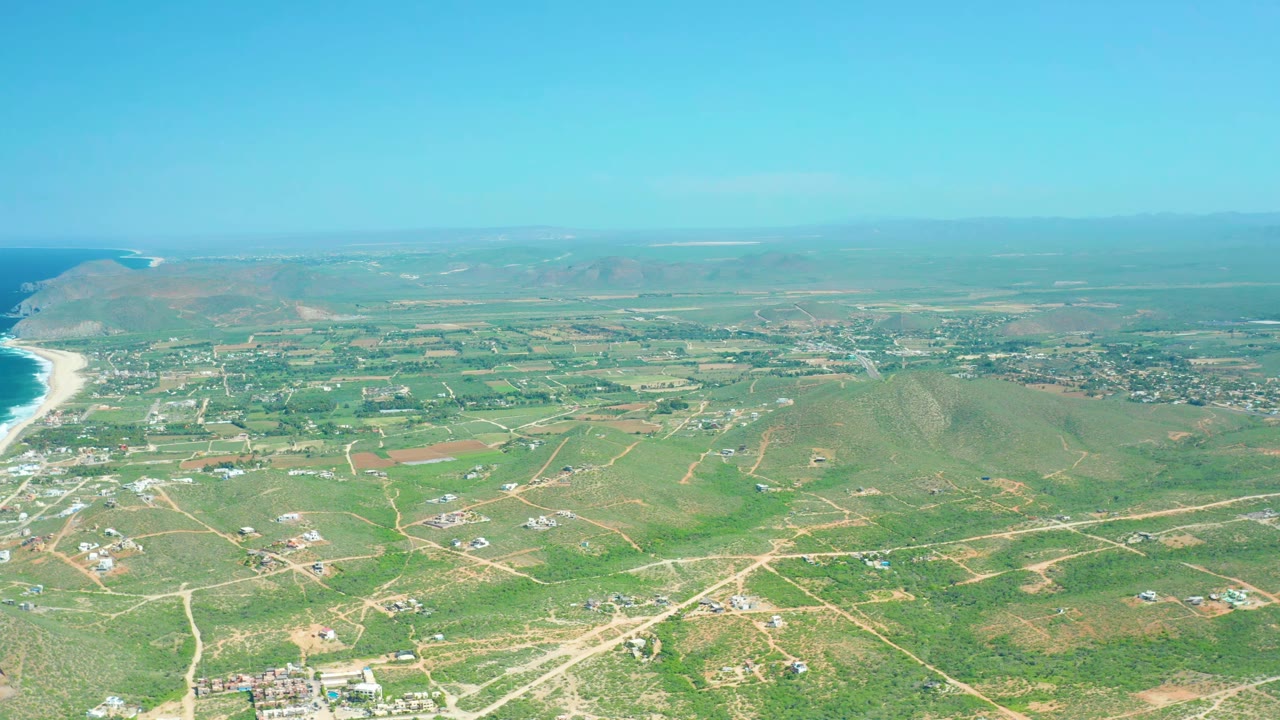 Coastal aerial with green fields, scattered buildings, beach, and horizon.