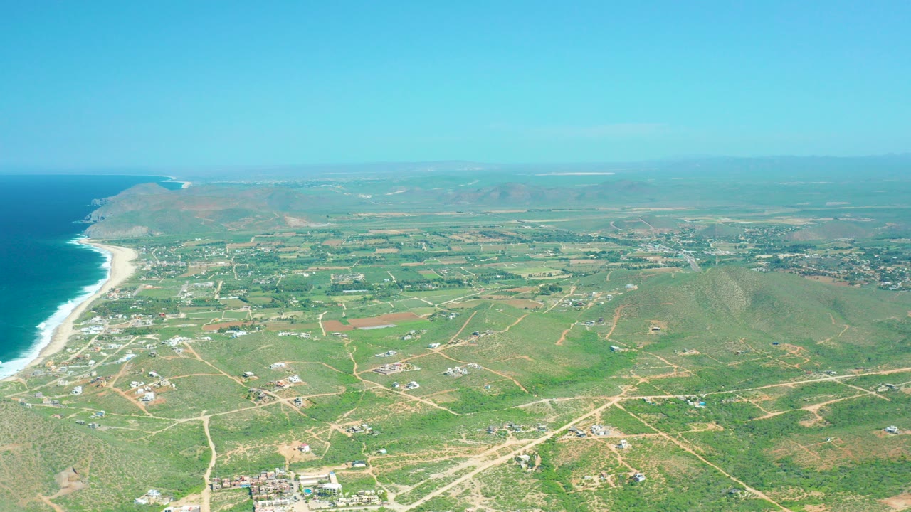 Wide aerial over Cerritos Beach in Baja California Sur, with blue ocean and white surf on the left, green fields and dirt roads inland, distant hills, and no visible people.