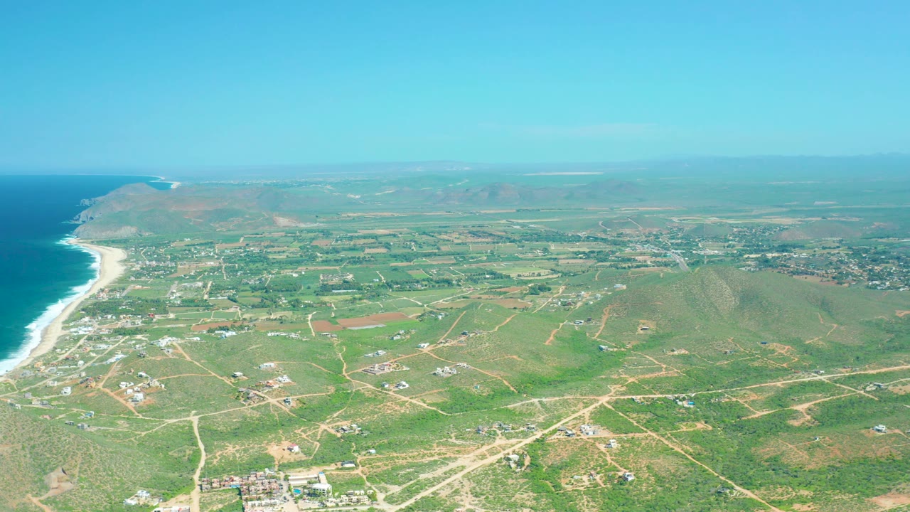 Cerritos Beach aerial view with the ocean and beach on the left, patchwork green farmland through the middle, light-colored roads, distant settlement, and no visible people.