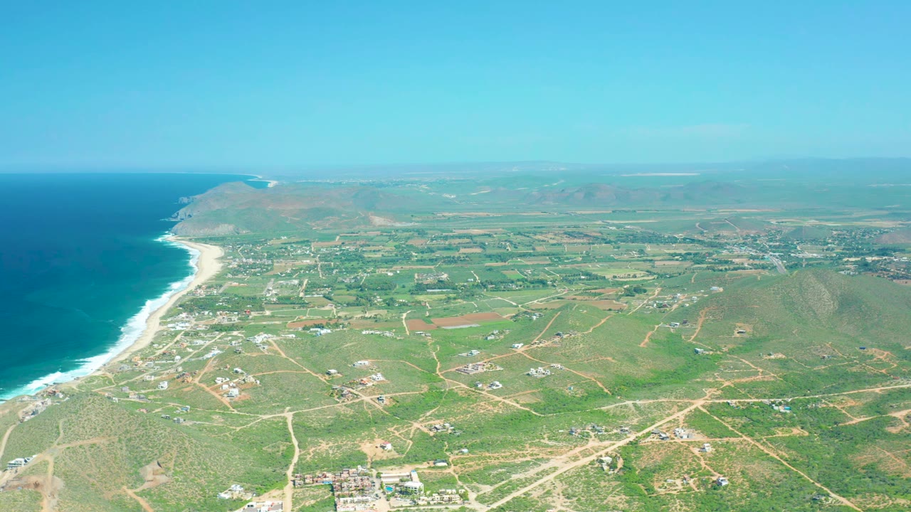 Aerial view of Cerritos Beach with the surf line curving along the left edge, green fields and pale dirt roads across the middle, scattered buildings near the lower frame, and no visible people.