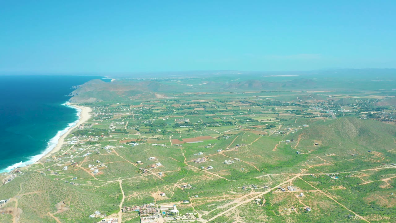 High drone frame over Cerritos Beach, Baja California Sur, with deep blue water on the left, a white surf band, inland farm-like fields, dirt tracks, low hills, and no visible people.