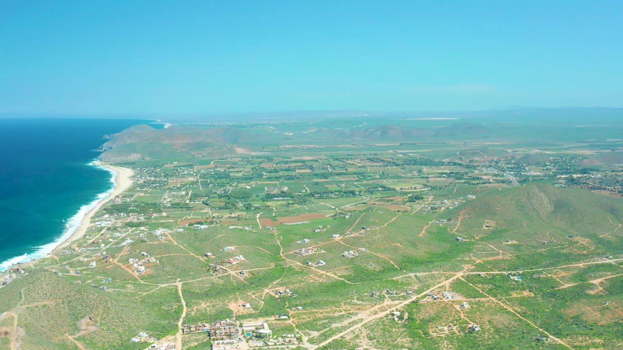 Mid-clip aerial frame showing Cerritos Beach shoreline at left, pale winding dirt roads through green and brown fields, scattered structures near the coast, low hills to the right, and no visible people.