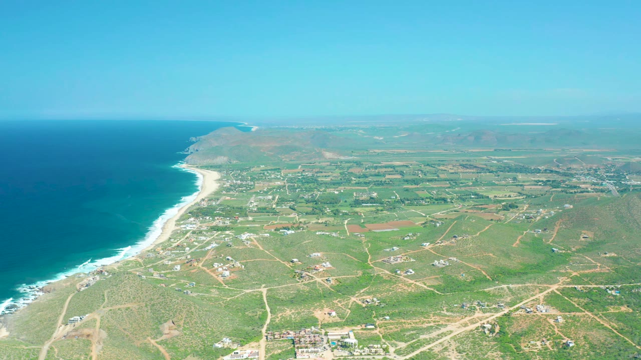 Wide drone frame of Cerritos Beach showing the surf line running diagonally below the ocean, a patchwork of green fields and dirt roads inland, distant hills, and no visible people.