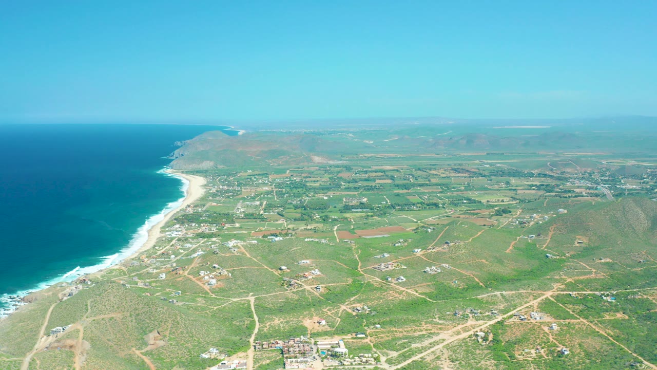 Aerial frame over Cerritos Beach with more of the beach curve visible, white surf bordering the ocean, inland fields divided by dirt roads, low buildings, hazy hills, and no visible people.