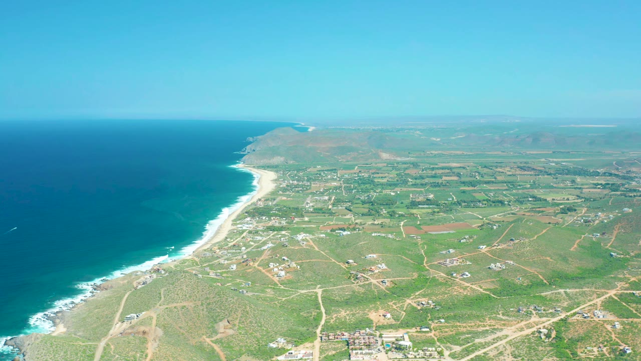 Wide aerial frame of Cerritos Beach with the ocean and surf line running along the left edge, farmland and dirt roads across the right side, distant hills on the horizon, and no visible people.