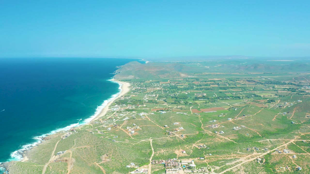Oblique aerial over Cerritos Beach showing the curved sandy coast, dark blue water, white surf bands, green inland plots, pale roads, scattered structures, and no visible people.