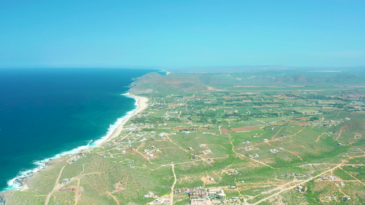 Mid-clip aerial view of Cerritos Beach with the coastline stretching into the distance, ocean at left, patchwork green fields and roads inland, low hills beyond, and no visible people.