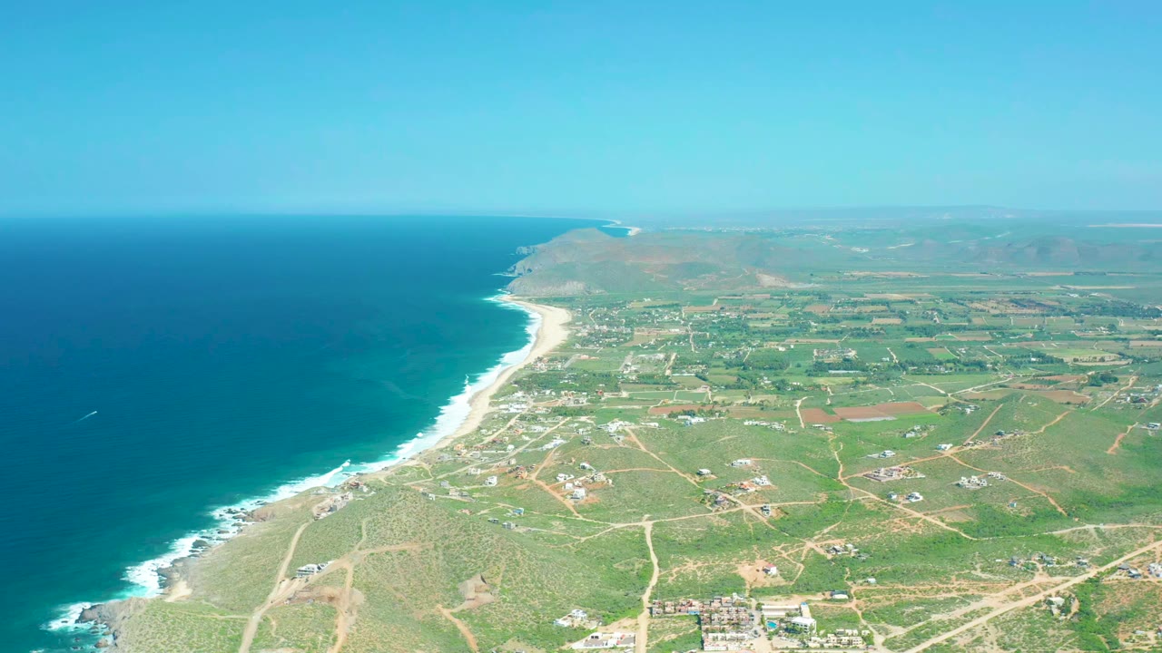 Aerial coastline with blue ocean, sandy surf line, fields, roads, and hills.