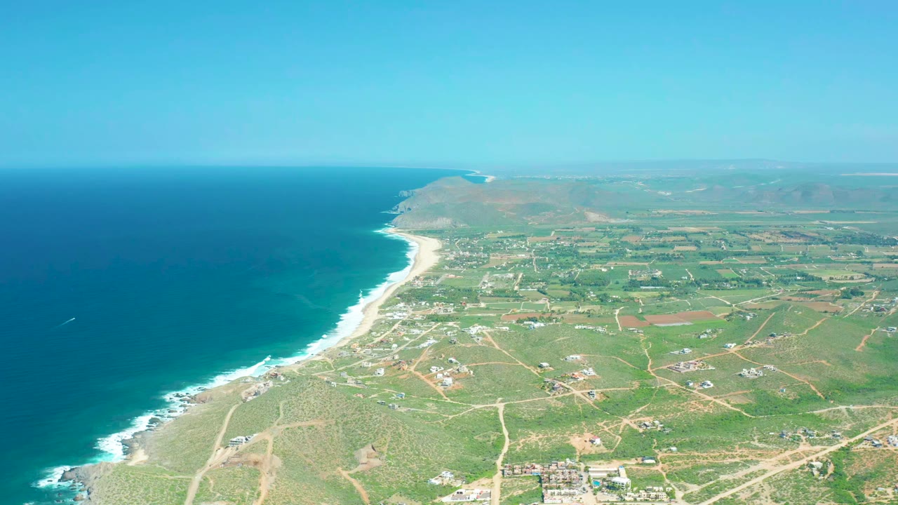 Aerial frame over Cerritos Beach showing the curving surf line, deep blue water, green inland parcels, pale dirt roads, scattered buildings, and no visible people.