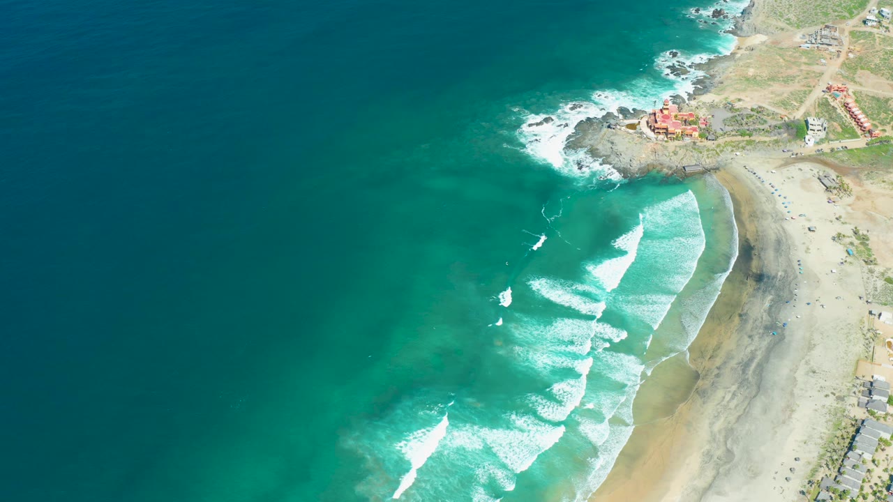 Turquoise ocean and whitecaps approach a sandy shore below a rocky coastline.