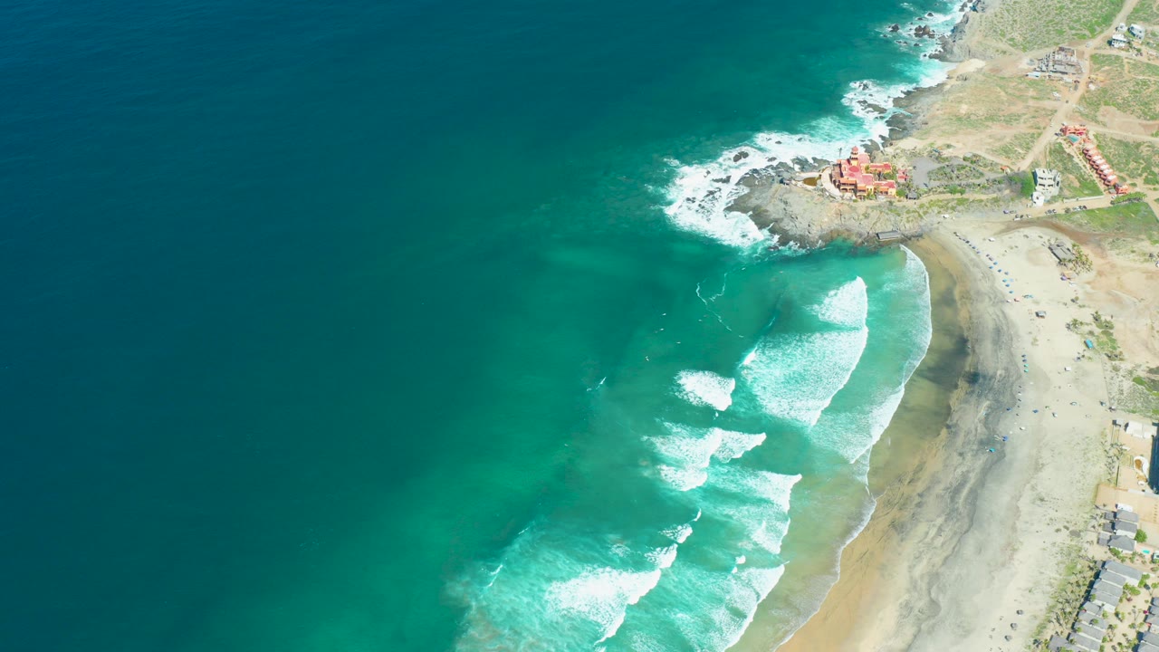 Opening aerial frame over Cerritos Beach in Baja California Sur, with deep blue water at left, white surf lines reaching sand at right, rocky coast and small structures near the top, and no visible people.