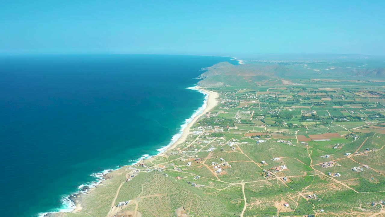 Aerial view over Cerritos Beach showing the surf line curving through the frame, inland green fields and dirt tracks to the right, distant hills, and no visible people.