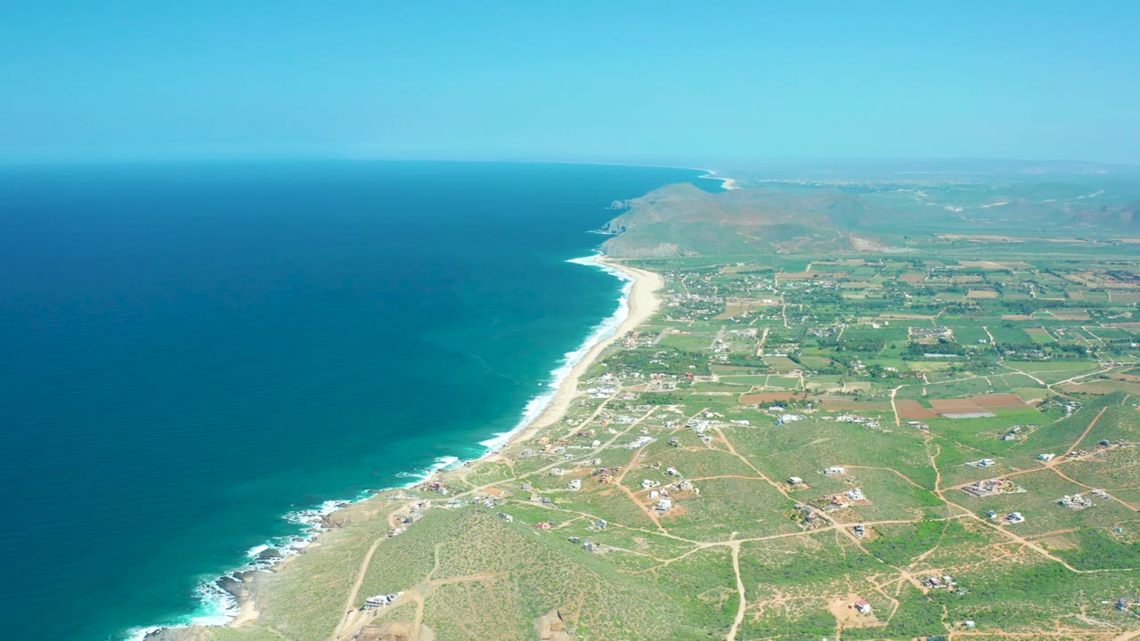 Wide aerial frame at Cerritos Beach with dark blue water filling the left side, pale beach and breaking waves along the coast, green rural plots inland, and no visible people.