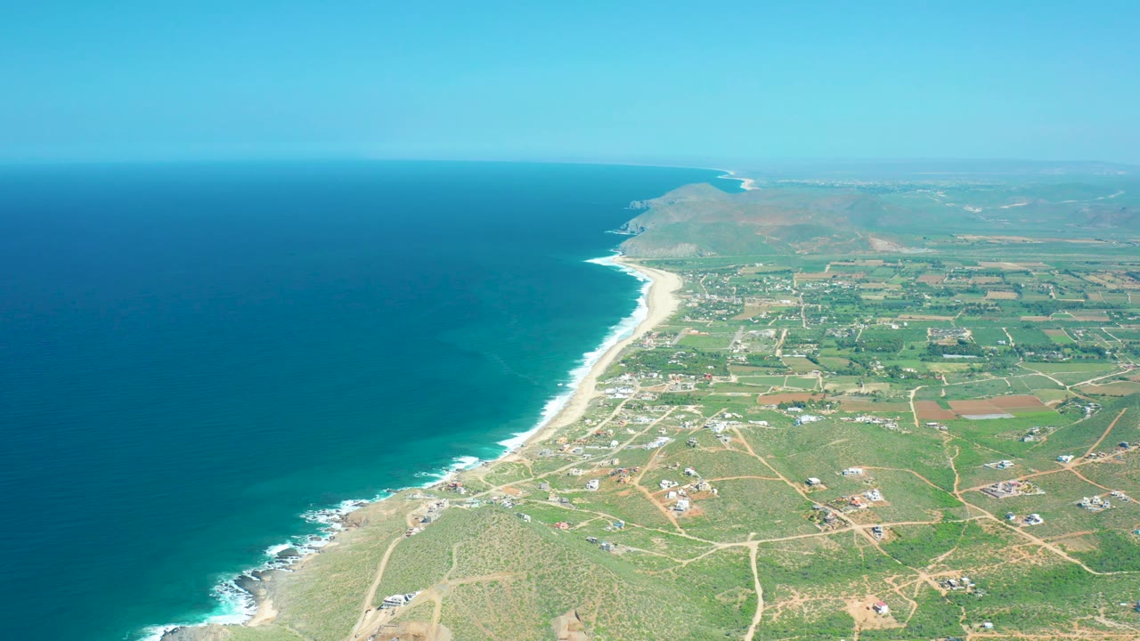 Mid-clip aerial view of Cerritos Beach with the shoreline running diagonally upward, white surf bands, patchwork green fields, scattered structures, and no visible people.