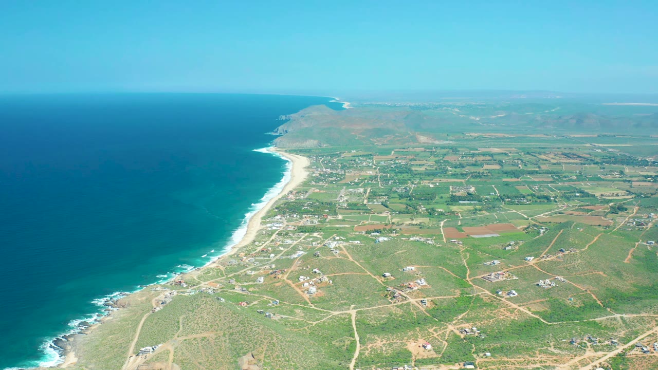 Aerial view at Cerritos Beach with the ocean filling the left side, waves marking the diagonal beach, patchwork farmland and small structures inland, a level horizon, and no visible people.