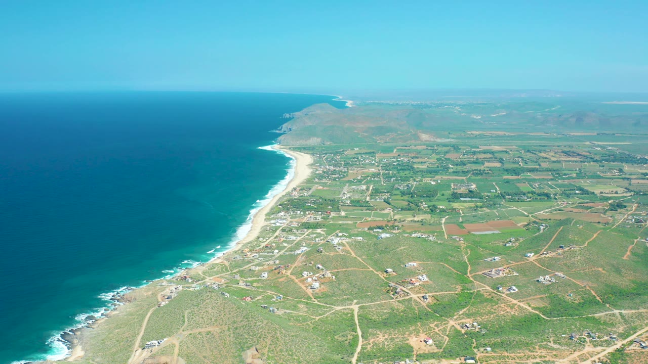 Cerritos Beach aerial frame showing darker blue water near the left edge, white waves along the beach, green inland plots, pale roads, scattered buildings, low hills in the distance, and no visible people.