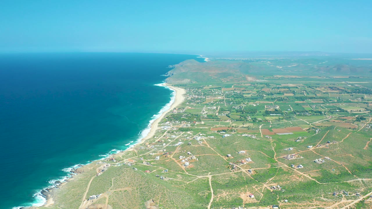 Mid-clip aerial over Cerritos Beach with a long curving shoreline, bright surf, turquoise and deep blue ocean, inland fields crossed by dirt roads, scattered buildings, hazy far hills, and no visible people.