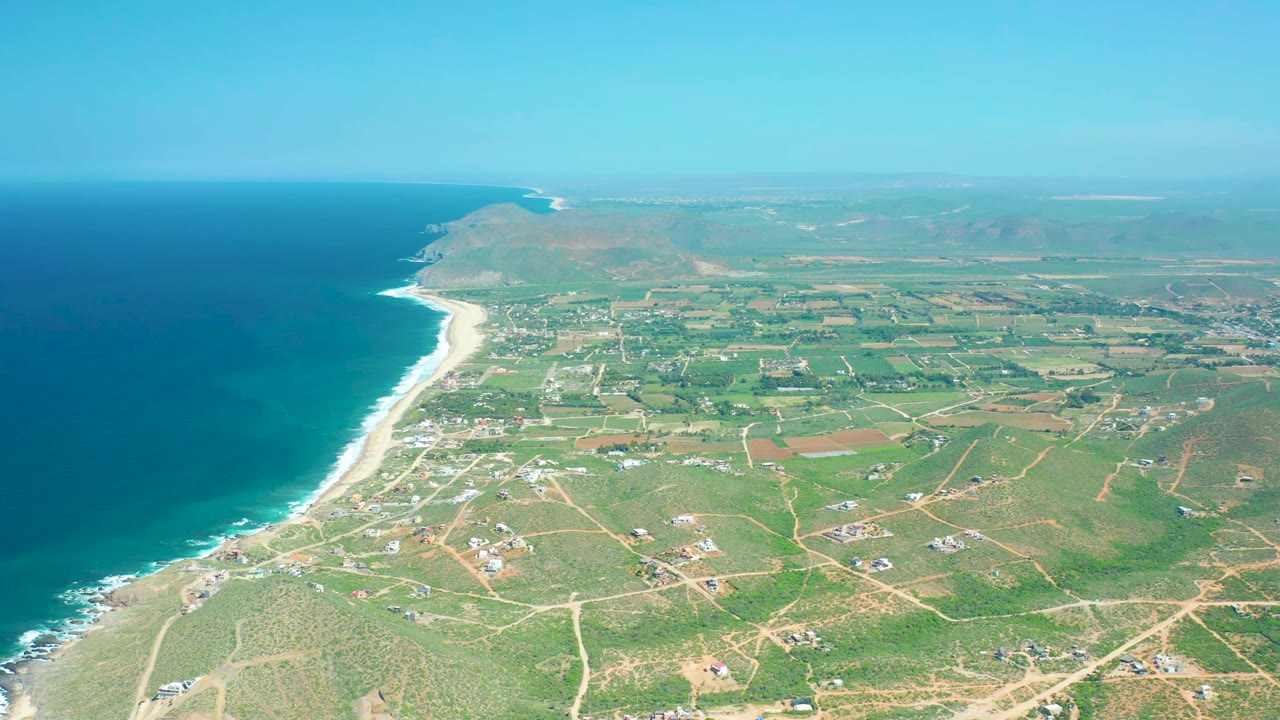 Aerial frame over Cerritos Beach showing the surf line curving up the coast, patchwork green and brown fields inland, light dirt roads, scattered buildings, distant low hills, and no visible people.