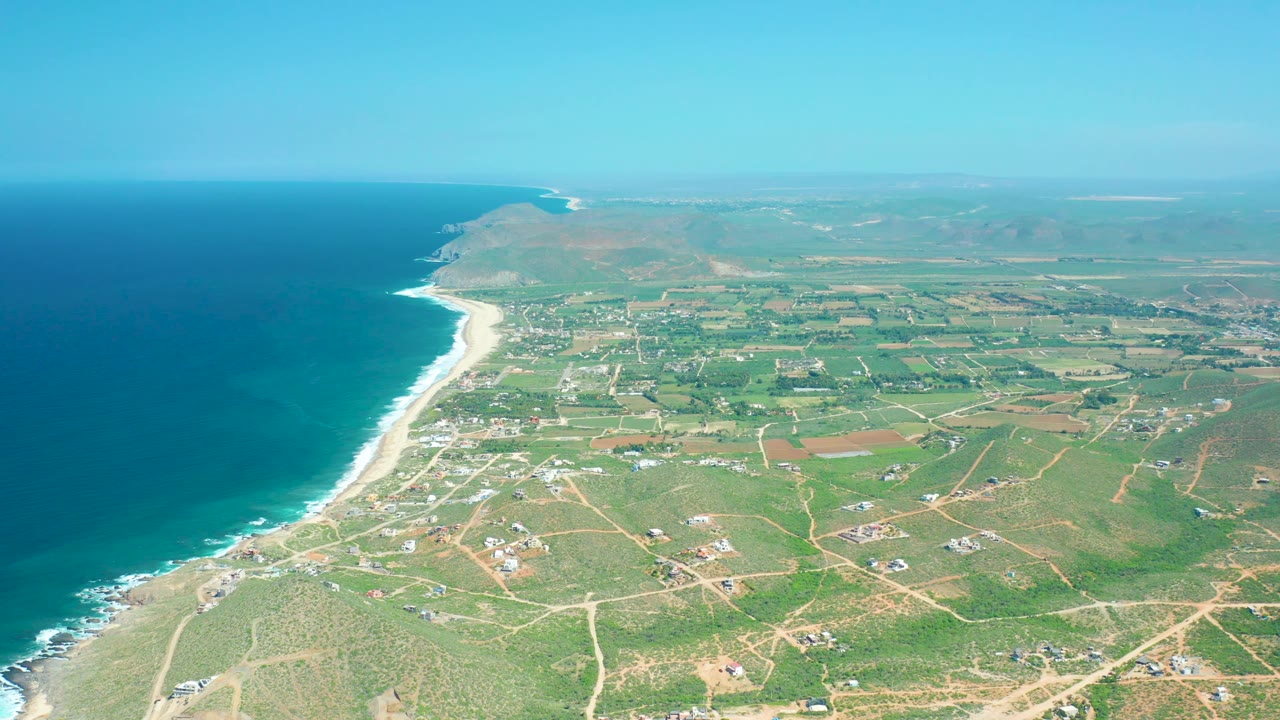 High aerial view of Cerritos Beach with deep blue water at left, white wave bands along the sand, inland green fields and roads across the right side, scattered structures, hazy horizon, and no visible people.