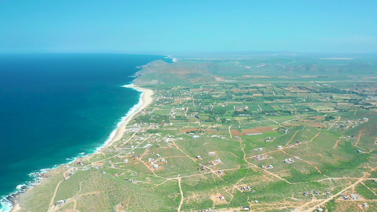 Drone-height view across Cerritos Beach, with active white surf along the left shoreline, pale sand, inland dirt-road grids, small buildings, green fields, distant hills, and no visible people.