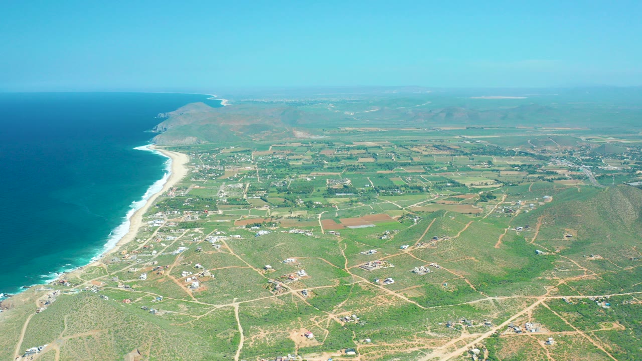 Aerial view of Cerritos Beach, Baja California Sur, with deep blue ocean on the left, white surf along the sand, inland fields and dirt roads to the right, and no visible people.