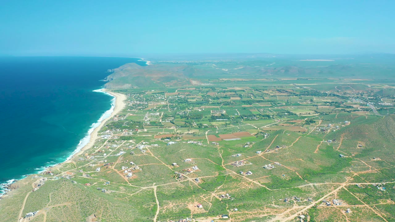 Aerial coastline with surf, farmland, dirt roads, and open sky.