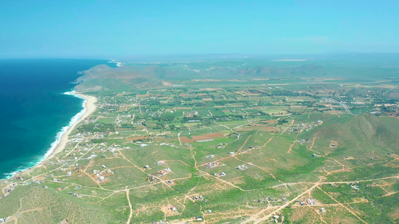 Wide aerial view over Cerritos Beach, Baja California Sur, with blue ocean at left, a sandy surf line, green fields, dirt roads, scattered buildings, and no visible people.