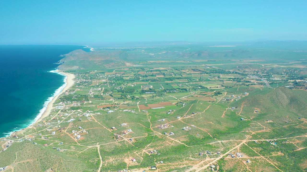 High aerial view of Cerritos Beach, Baja California Sur, showing turquoise-blue water, a pale beach edge, inland farmland, scattered structures, hazy distance, and no visible people.