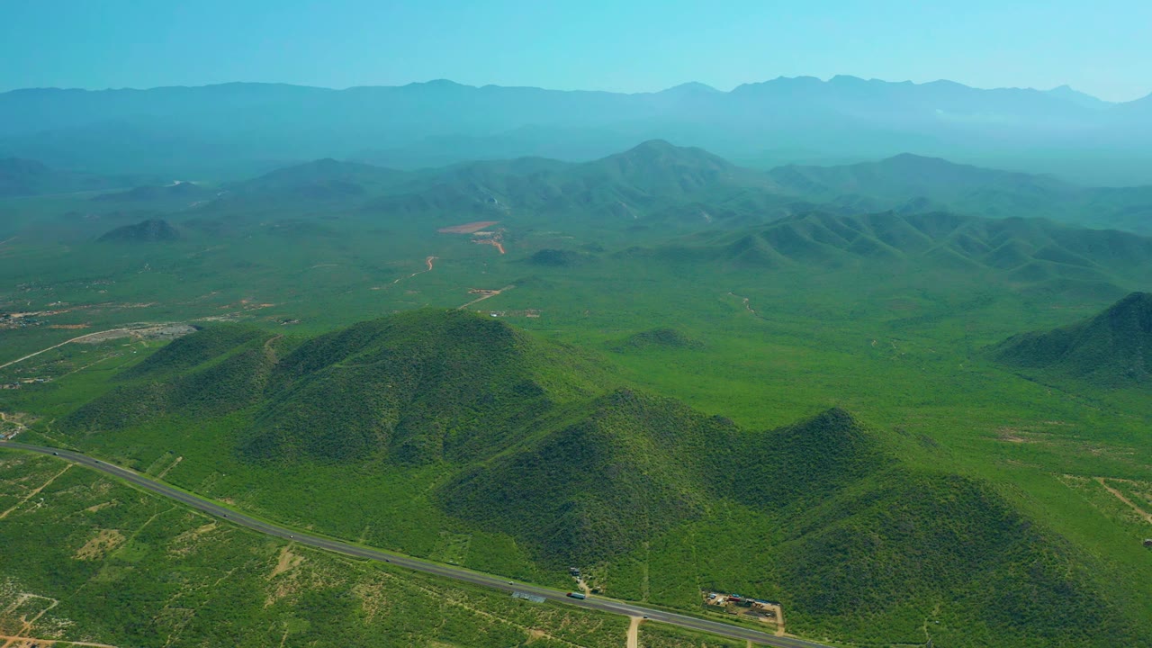 Aerial frame near Cerritos Beach with small vehicles on a paved road, rounded green hills in the foreground, layered mountains in the distance, and no visible people.