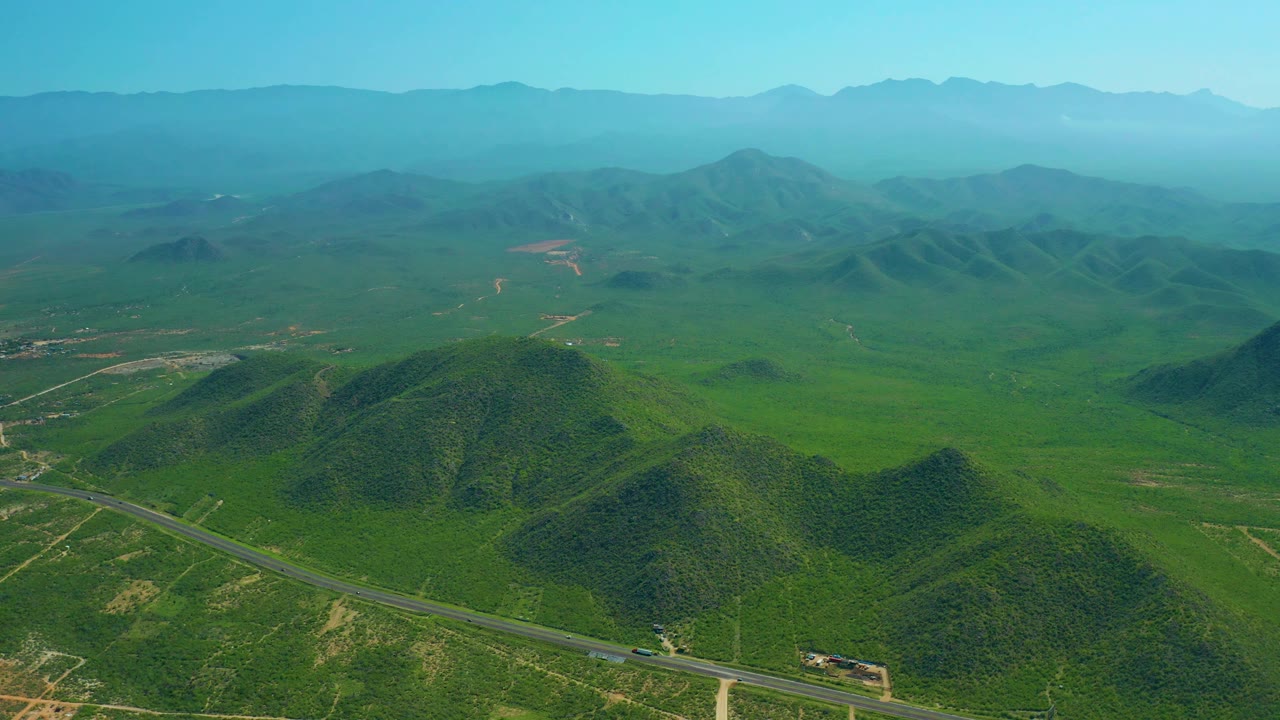 Aerial view in Cerritos Beach, Baja California Sur, with the road angling across the lower frame, green hills in the center, distant ridges, and no visible people.